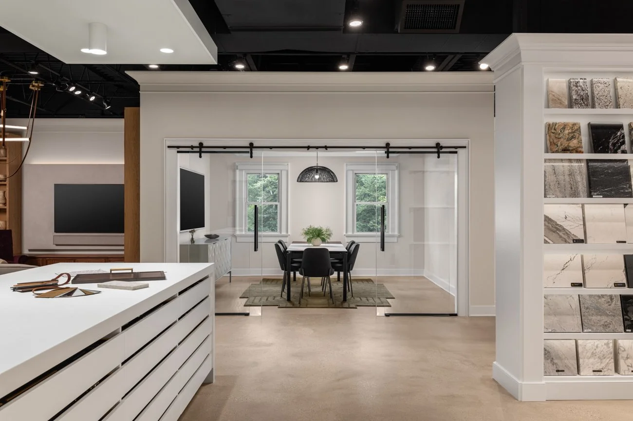 Showroom interior with a dining area enclosed by glass sliding doors, a black dining table with chairs, and large tile samples on the walls.