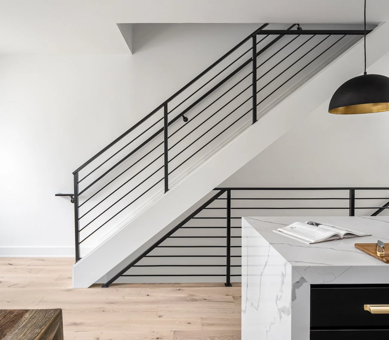 Interior view of a modern space with black metal railing on a staircase, white walls, a marble countertop, and a black and gold hanging lamp.