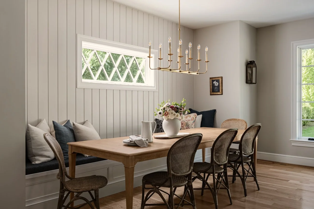 A dining room with a wooden table, six woven chairs, a window with diamond-patterned glass, a built-in window seat with pillows, a flower vase with flowers, a chandelier, and sunlight coming through the windows.
