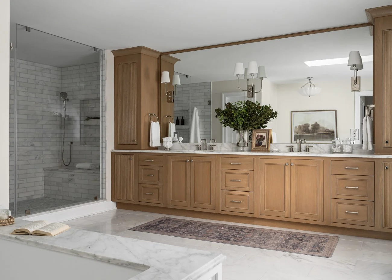 A spacious bathroom with a wooden vanity, large mirror, and a glass-enclosed shower with white subway tiles. Decor includes a large green plant, framed artwork, and bedside lamps, with marble countertops and a beige rug on a tiled floor.