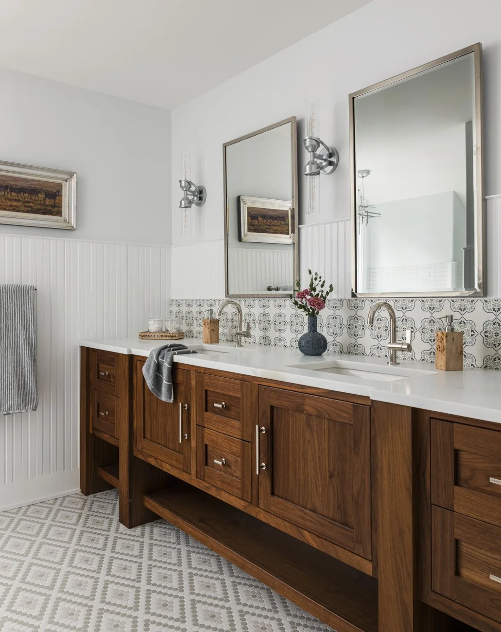Bathroom with wooden vanity and white countertop, two large mirrors, decorative tiles behind sinks, and vases with flowers.