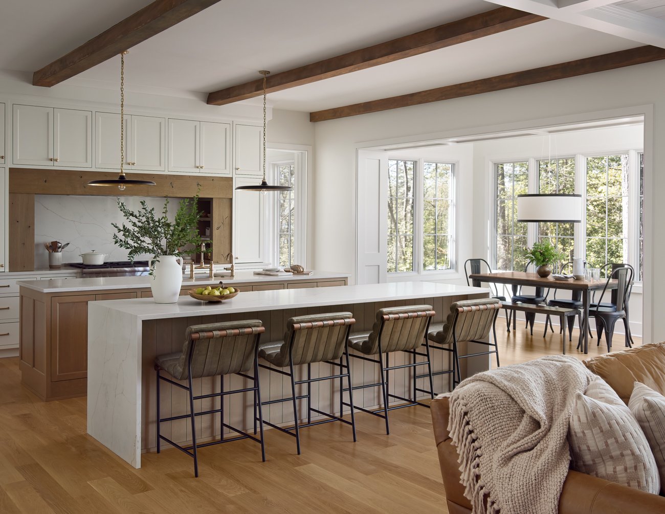 Open-concept kitchen and dining area with wooden floors, white cabinets, a large kitchen island with bar stools, and a dining table with chairs near bay windows, decorated with plants and a modern lamp.