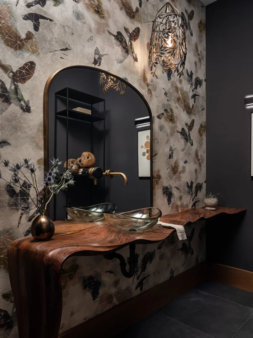 A bathroom vanity with a live edge wooden countertop, black vessel sinks, and a round mirror with a black frame. The wall features a butterfly and flower patterned wallpaper, and there are decorative items including a bronze vase with flowers, skull 