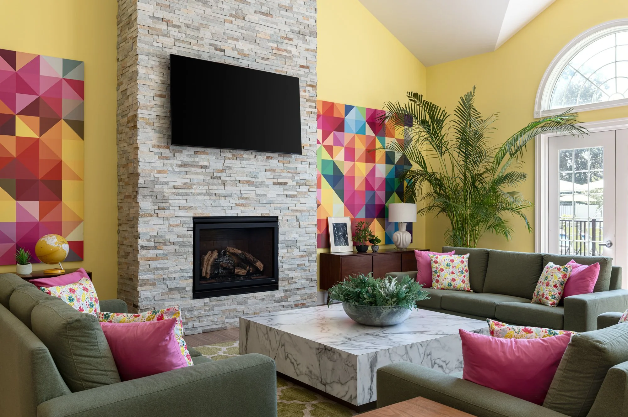 Living room with a stone fireplace, mounted flat-screen TV, colorful abstract wall art, a marble coffee table, sea-green sofas with pink and floral pillows, a wooden sideboard with picture frames, a lamp, and large indoor plants near a glass door wit