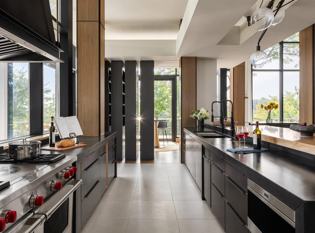 Modern kitchen with black cabinets, stainless steel appliances, and large windows with a view of greenery, decorated with sunflowers and a bottle of red wine.