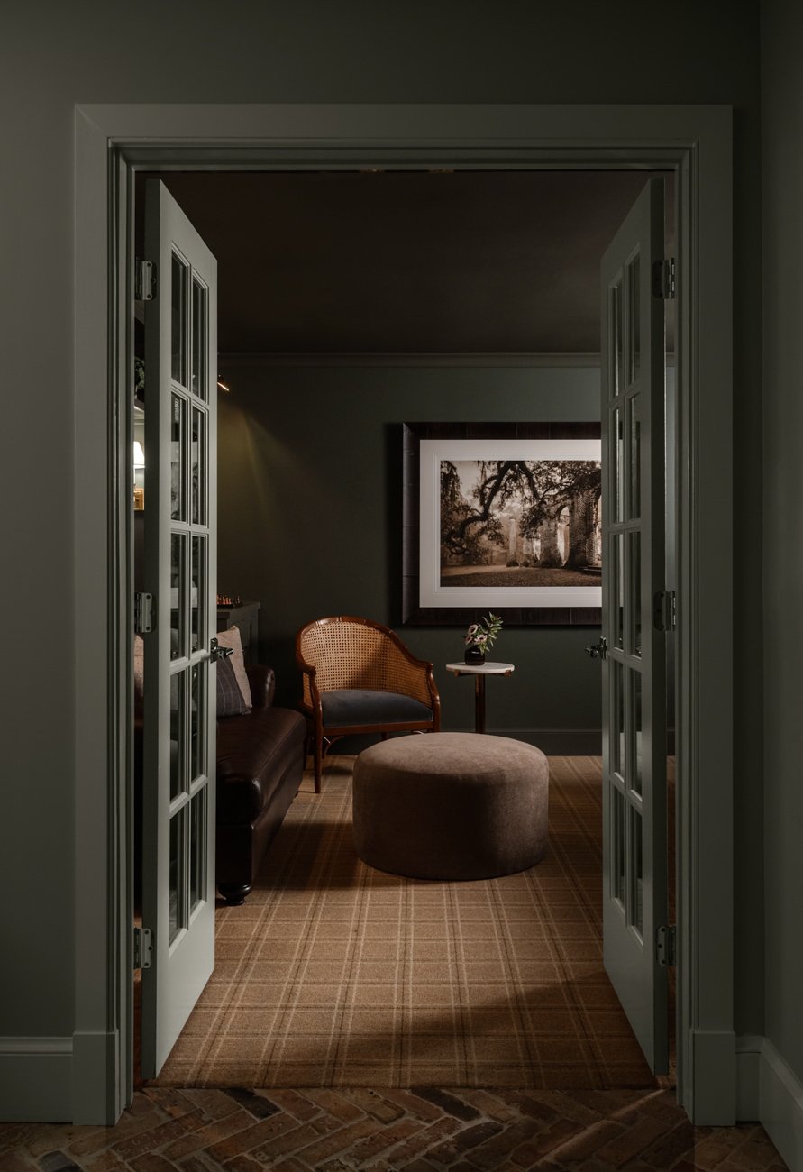 View of a cozy sitting room through French doors with a dark green wall, a framed black-and-white photograph of trees, a cane-back armchair, an upholstered ottoman, a small side table with a flower vase, and a leather sofa.