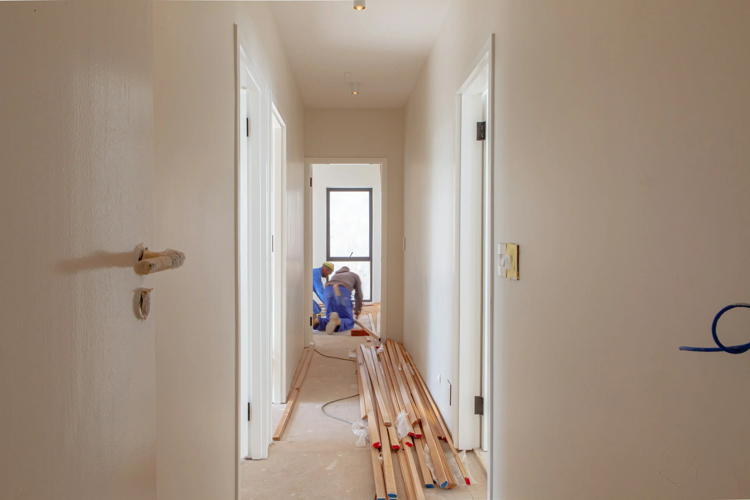 A hallway under construction with wooden flooring materials laid out on the floor. Workers are installing the flooring near a door at the end of the hallway. The walls are painted white, and there is a window at the far end letting in natural light.