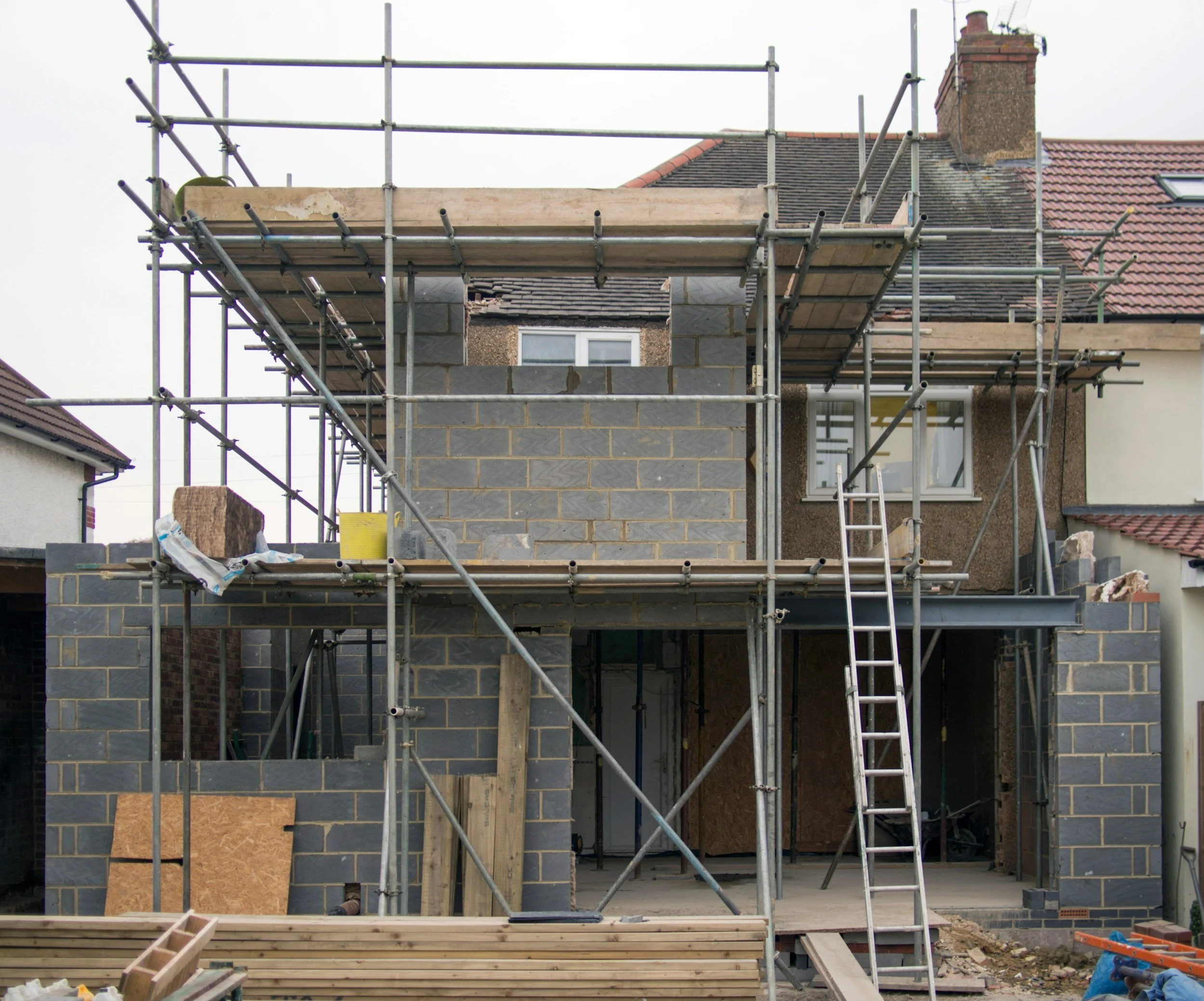 Construction site with scaffolding around a partially built brick house. Wood planks and construction materials are visible at ground level.