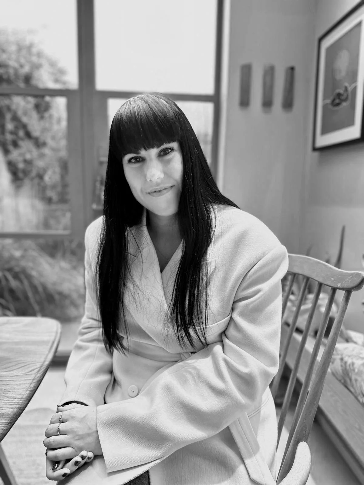 A woman with long dark hair and bangs sitting at a wooden table in a room with large windows, some framed artwork on the wall, and a garden outside.
