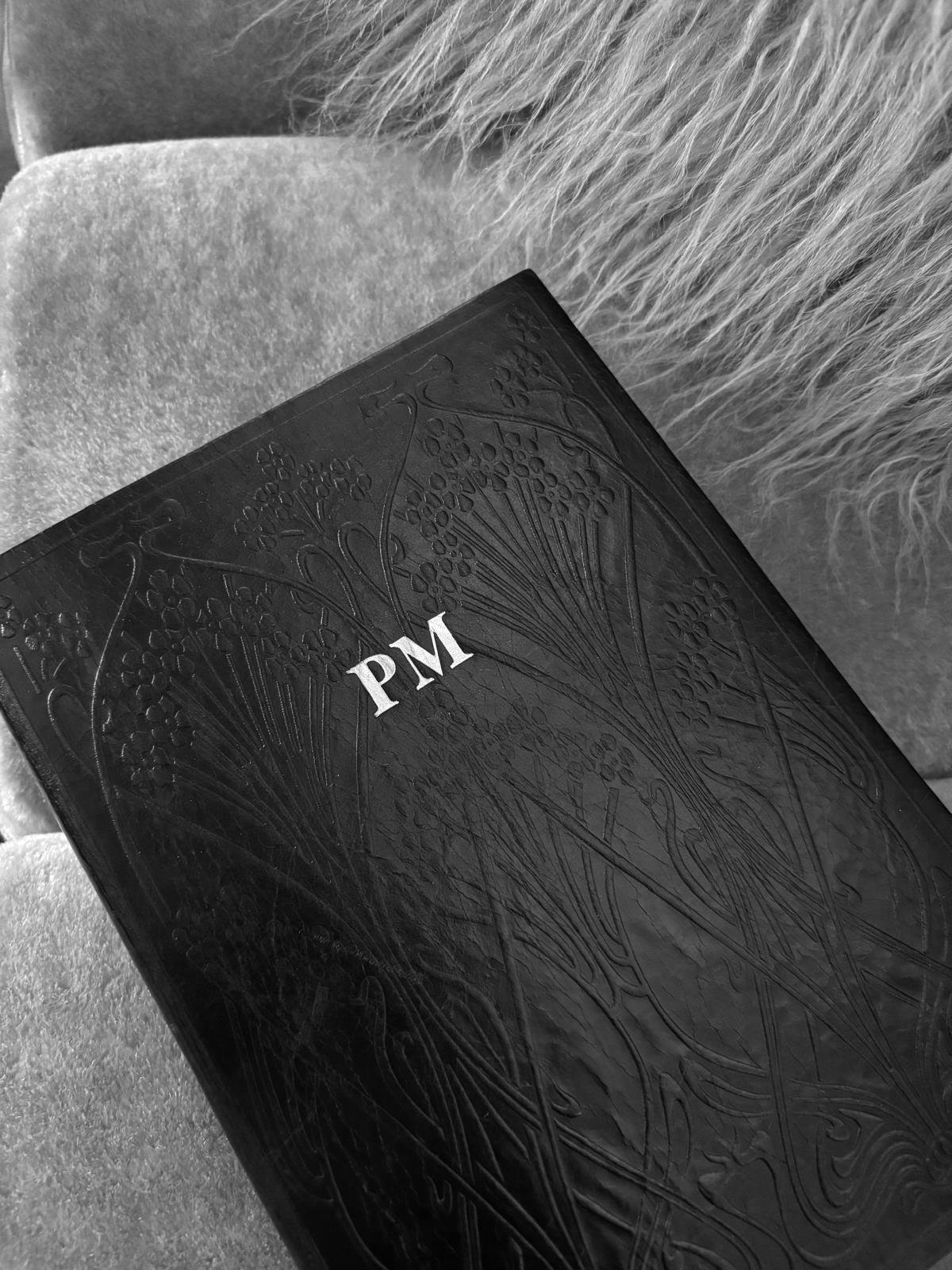 A black leather-bound book with engraved designs and the letters 'PM' on the cover, resting on a textured surface.