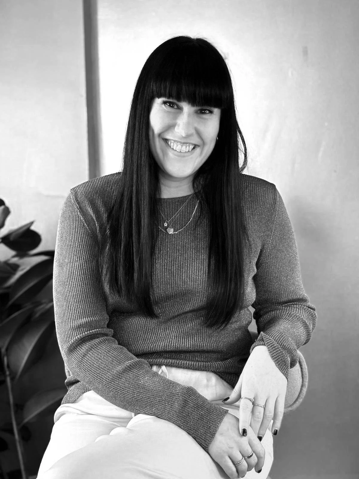 Black and white photograph of a smiling woman with long dark hair and bangs, wearing a sweater and layered necklaces, sitting indoors next to a plant.