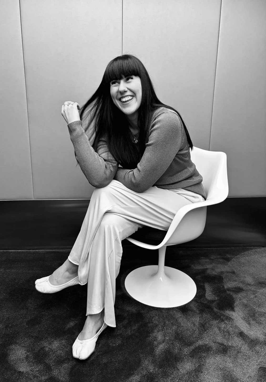 A woman with long dark hair and bangs, smiling happily while sitting on a modern white chair against a plain wall, dressed in a long-sleeve top, light-colored pants, and flat shoes.