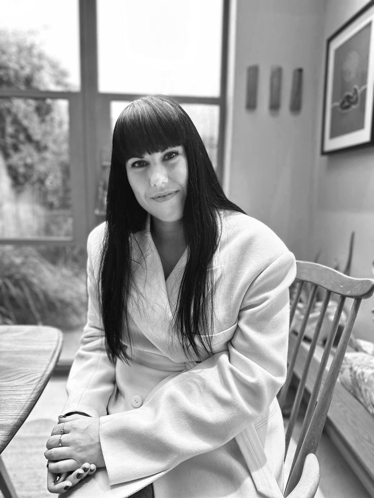 A woman with long dark hair and bangs, wearing a light-colored coat, sitting at a wooden table in a room with large windows, art on the wall, and outdoor greenery visible through the windows.