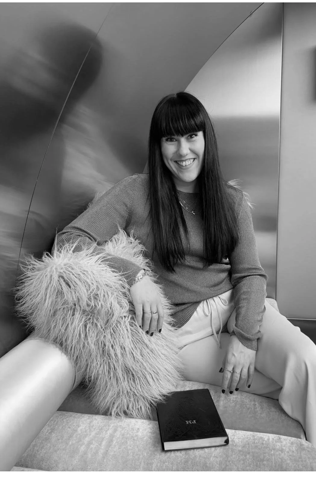 A woman with long dark hair and bangs smiling while sitting on a lounge with a furry dog. She is wearing a sweater and light-colored pants. There is a notebook on the lounge in front of her.