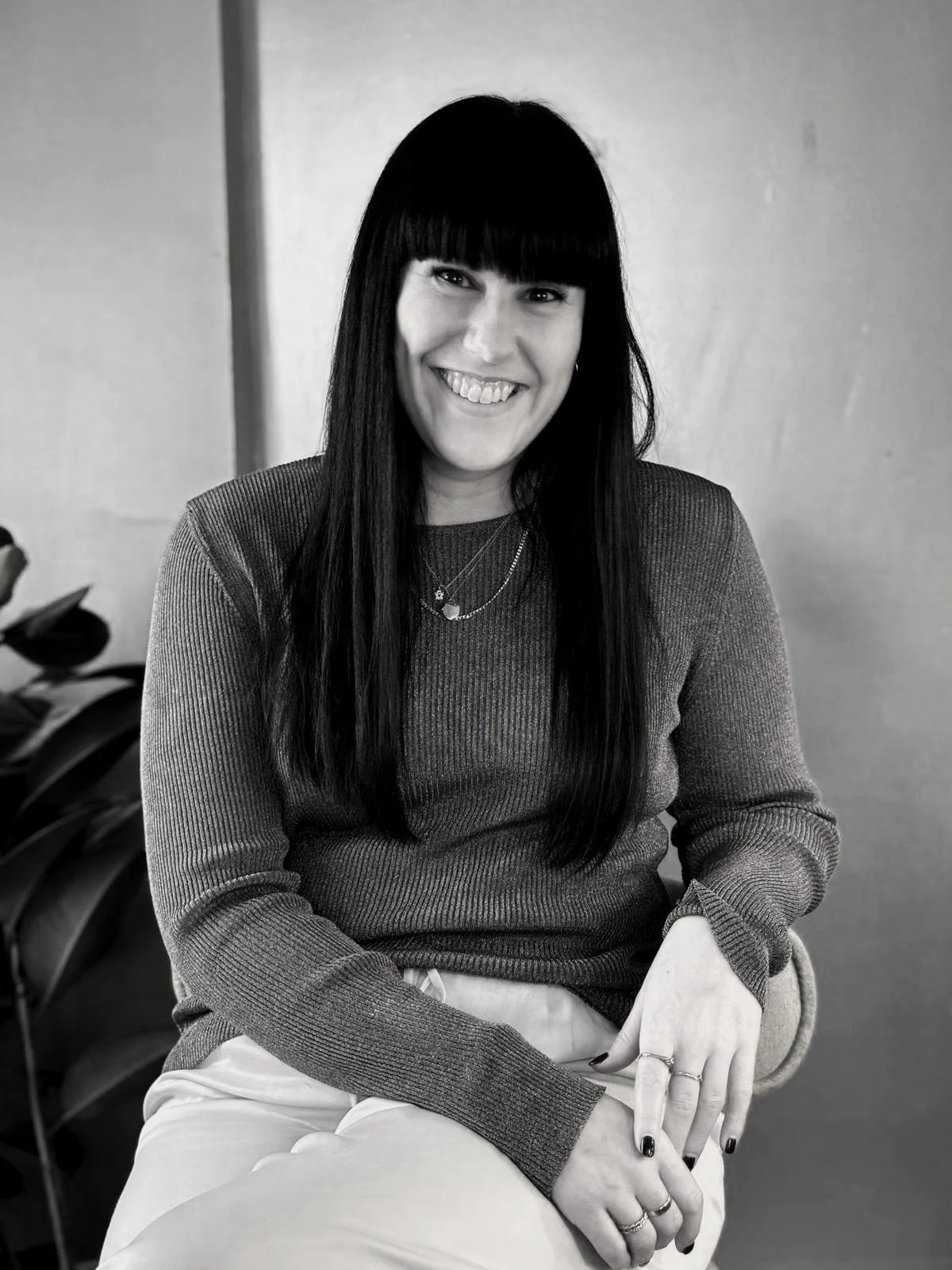 A woman with long dark hair and bangs, smiling, sitting on a chair indoors.