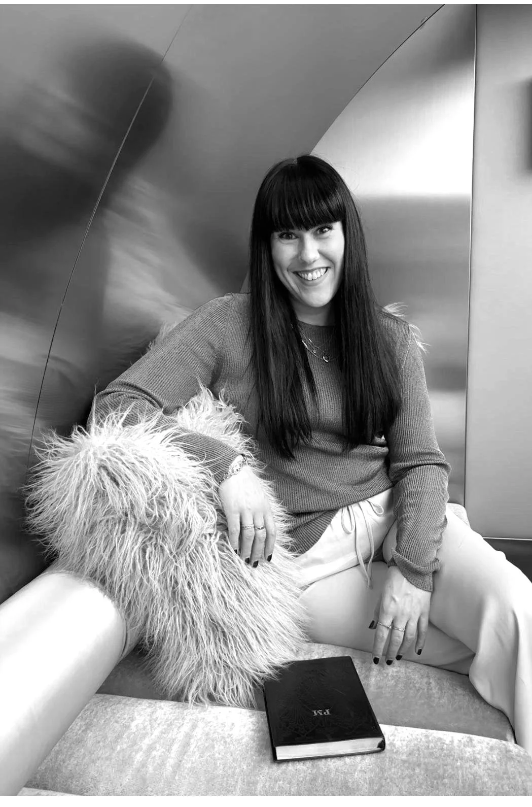 A woman with long dark hair and bangs sitting on a sofa next to a person in a fluffy coat. She is smiling at the camera, wearing a long-sleeve top and light-colored pants. There is a closed black book on her lap.