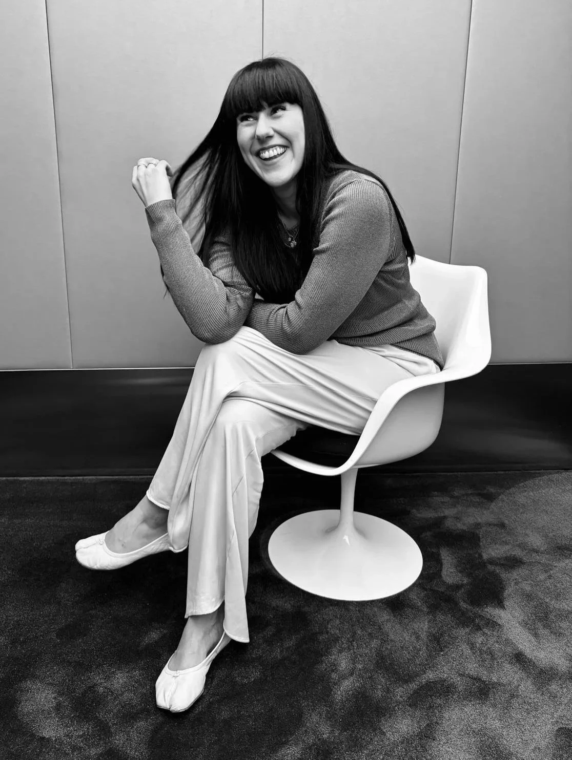 A woman with long dark hair and bangs sitting on a modern white chair, smiling and touching her hair in a casual pose, in a room with plain walls and dark carpet.
