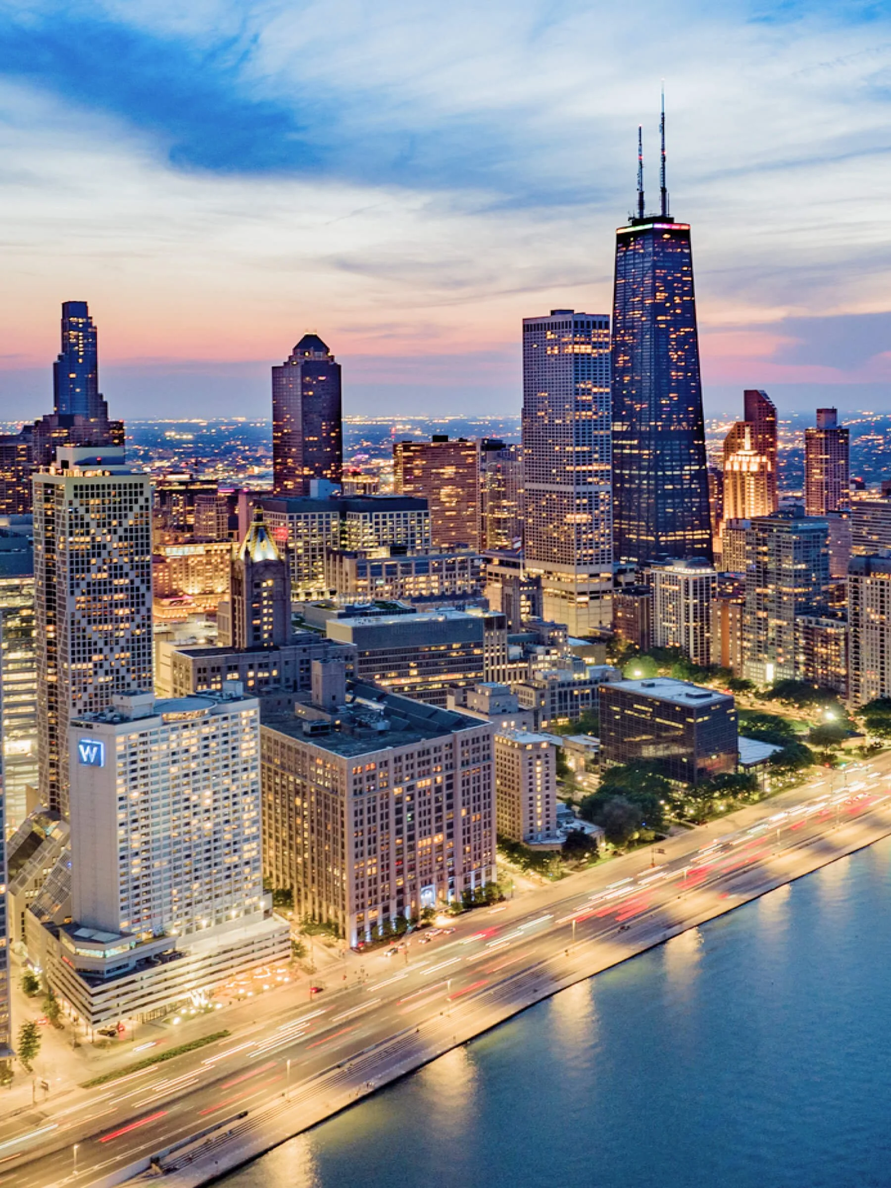 Aerial view of Chicago skyline at dusk with illuminated skyscrapers, including the John Hancock Center, along Lake Michigan.