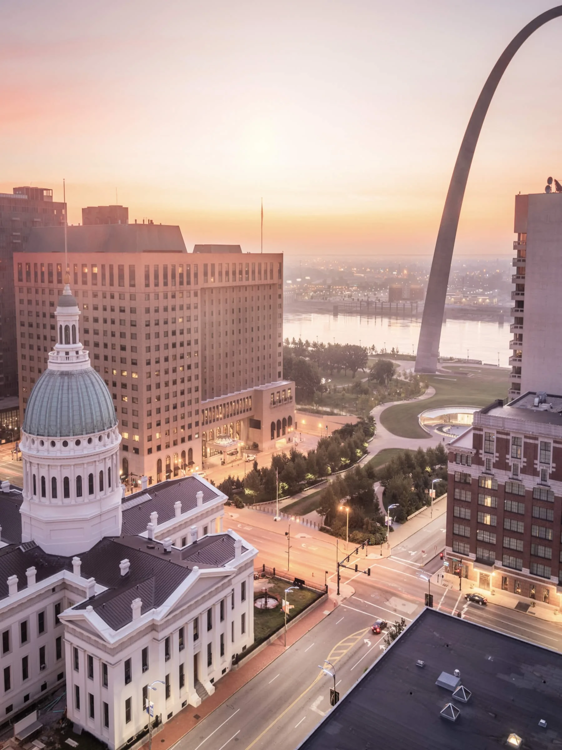 Cityscape of downtown St. Louis, Missouri during sunset, featuring the Gateway Arch, historic buildings, and the Mississippi River in the background.