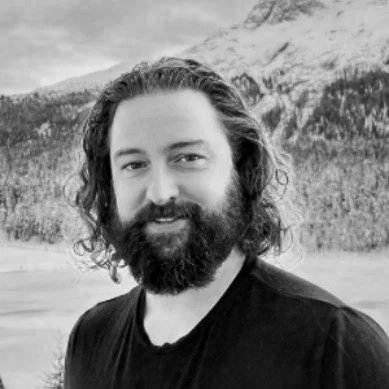 A man with long curly hair and a beard, wearing a black shirt, outdoors with a mountainous background.