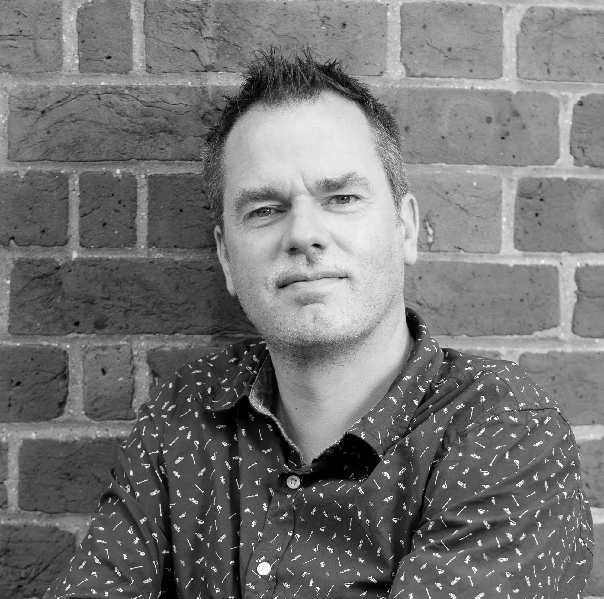 Black and white portrait of a man with short, spiky hair, wearing a patterned shirt, standing in front of a brick wall.