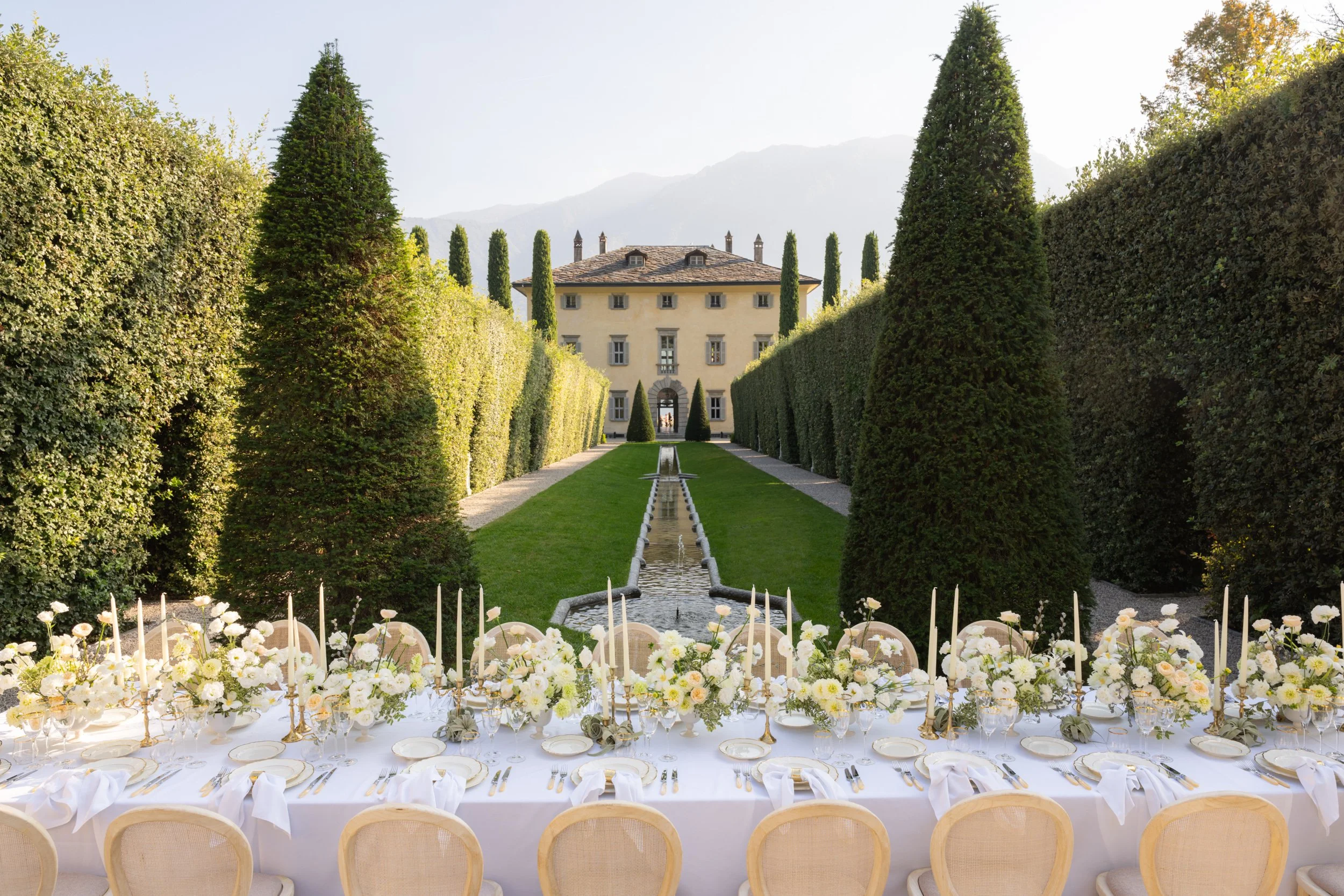 Long outdoor banquet table set with white tablecloth, white plates, gold cutlery, white napkins, floral arrangements, and candles, overlooking a formal garden and grand villa, with mountains in the background.