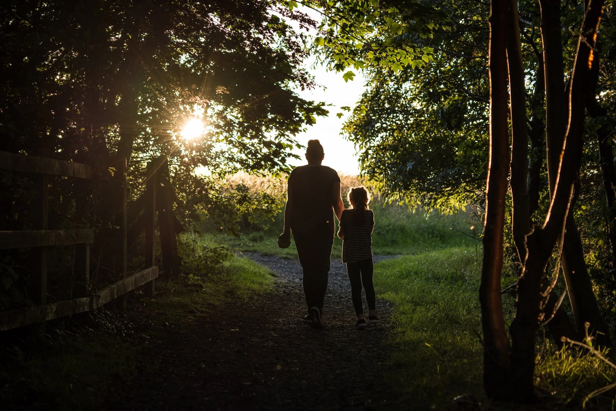 A person and a child walking on a trail in a forest during sunset with sunlight shining through trees.
