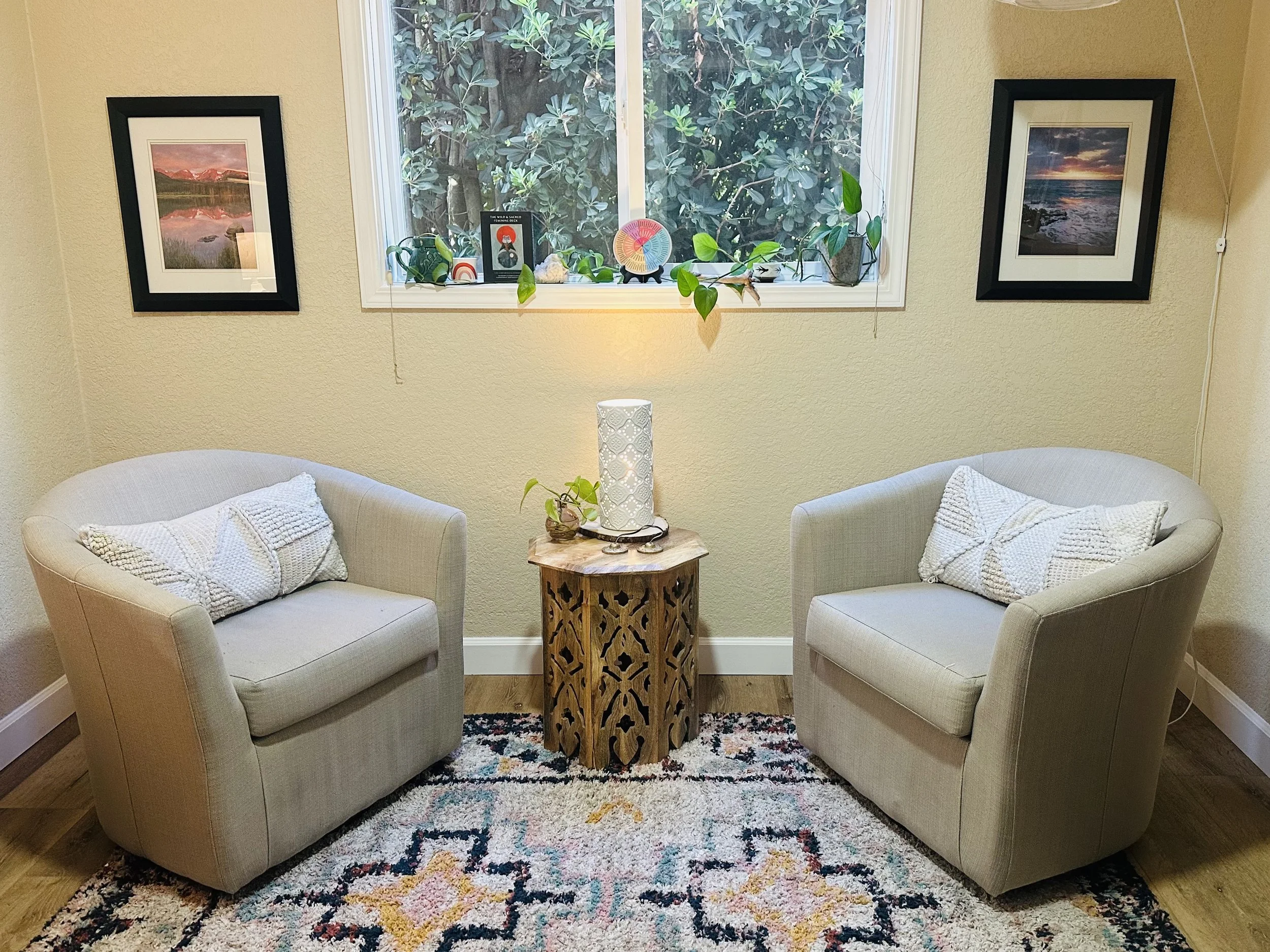 A cozy living room corner with two beige armchairs, a small wooden side table with a white lamp, decorative pillows, and a patterned area rug. A window behind them shows green foliage outside and displays framed photographs and decorative items on the windowsill.