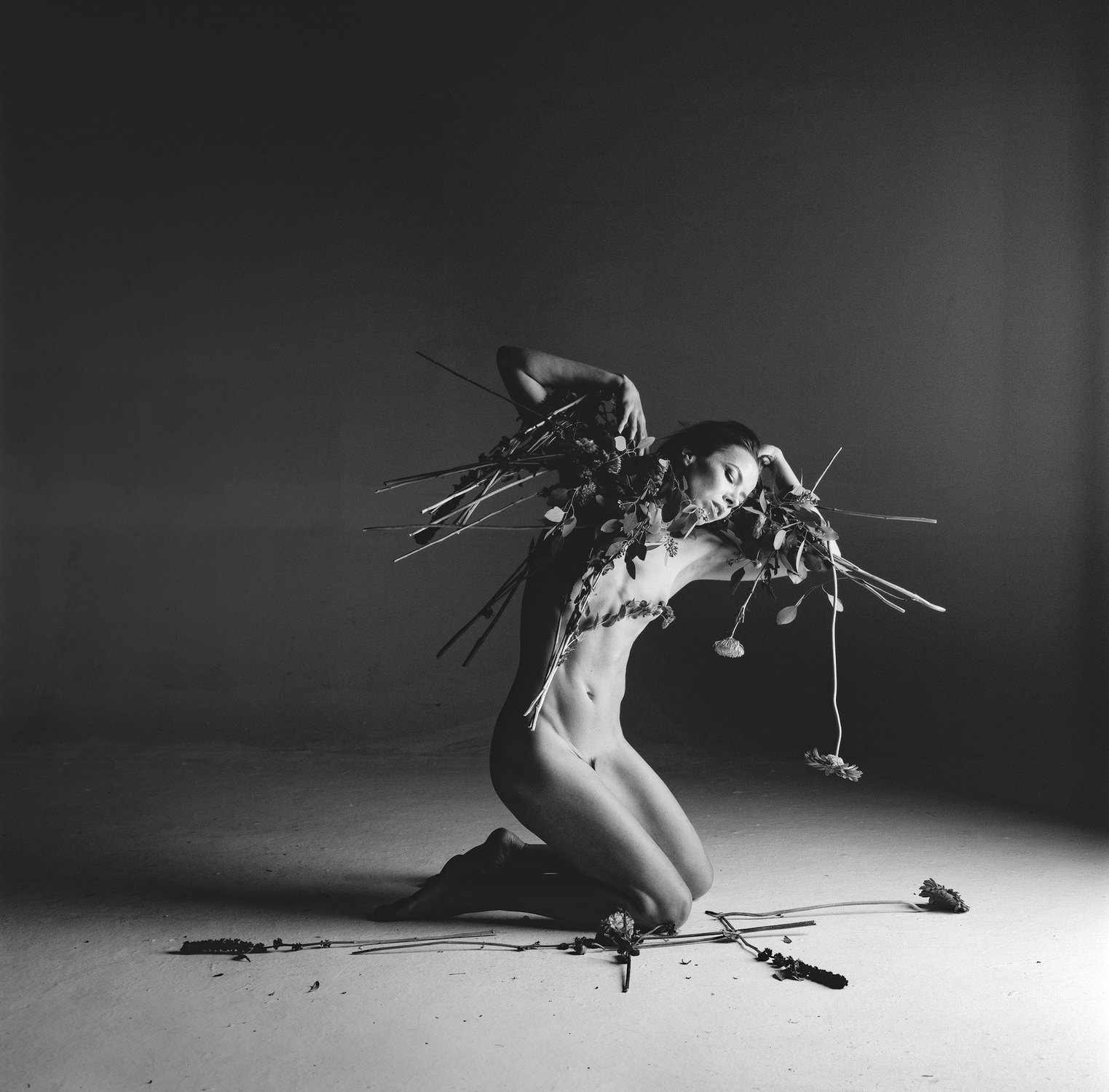 Black and white photo of a person kneeling with flowers and leaves covering their body, lit dramatically from the right.