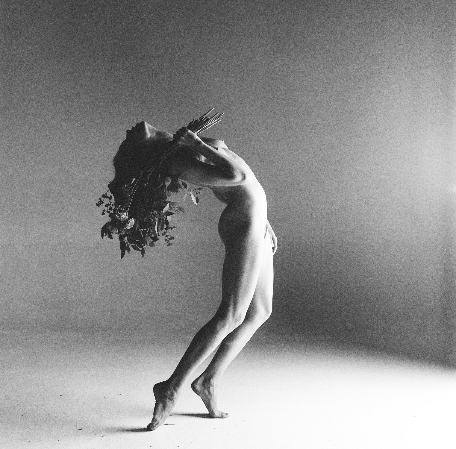 Black and white photo of a nude figure leaning back, holding a bouquet of flowers, with dramatic lighting and shadows.