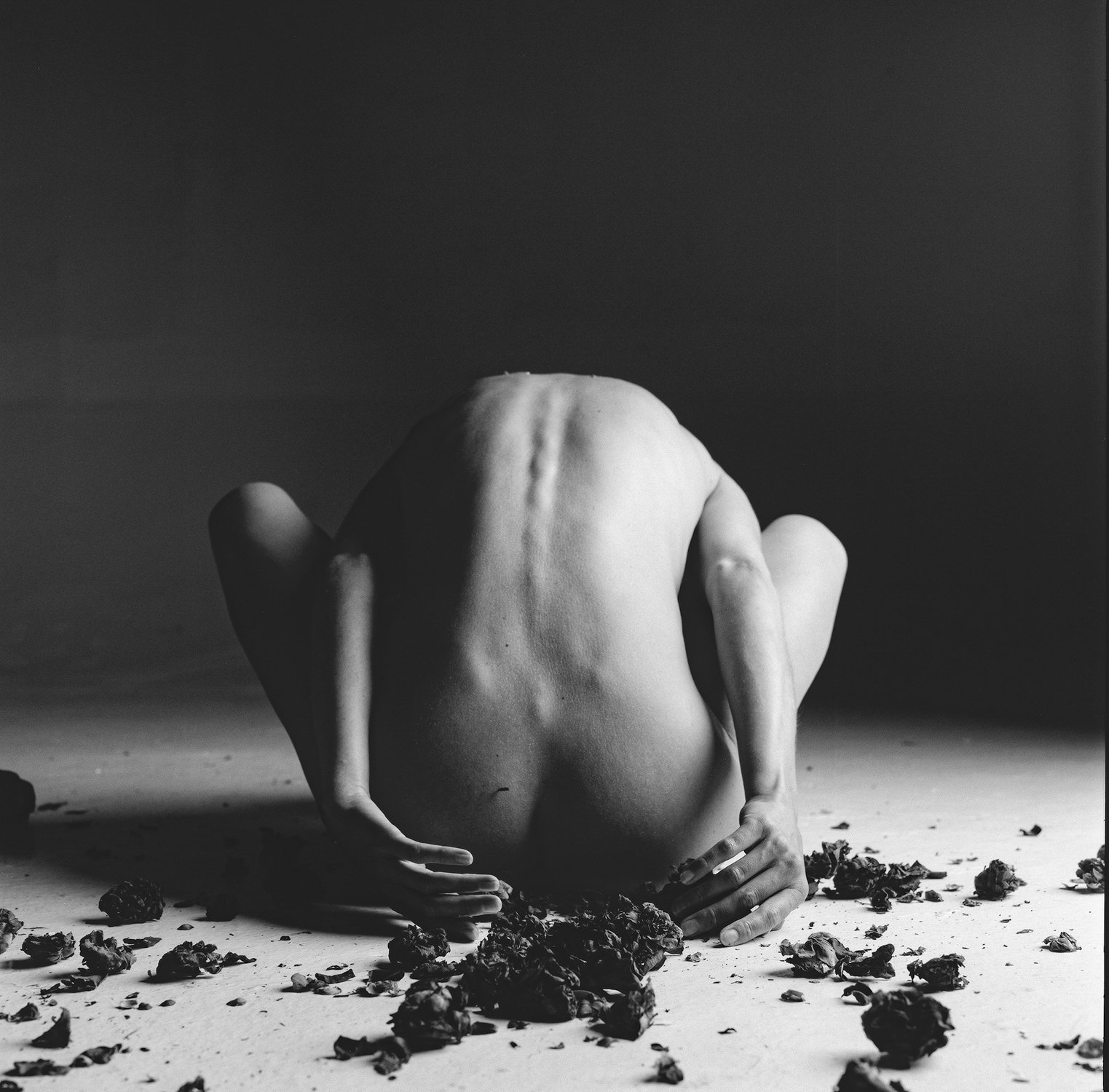 Black and white photo of a nude person crouched with their back facing the camera, surrounded by scattered dried leaves or debris on the ground. The room is dimly lit, emphasizing shadows and contours.