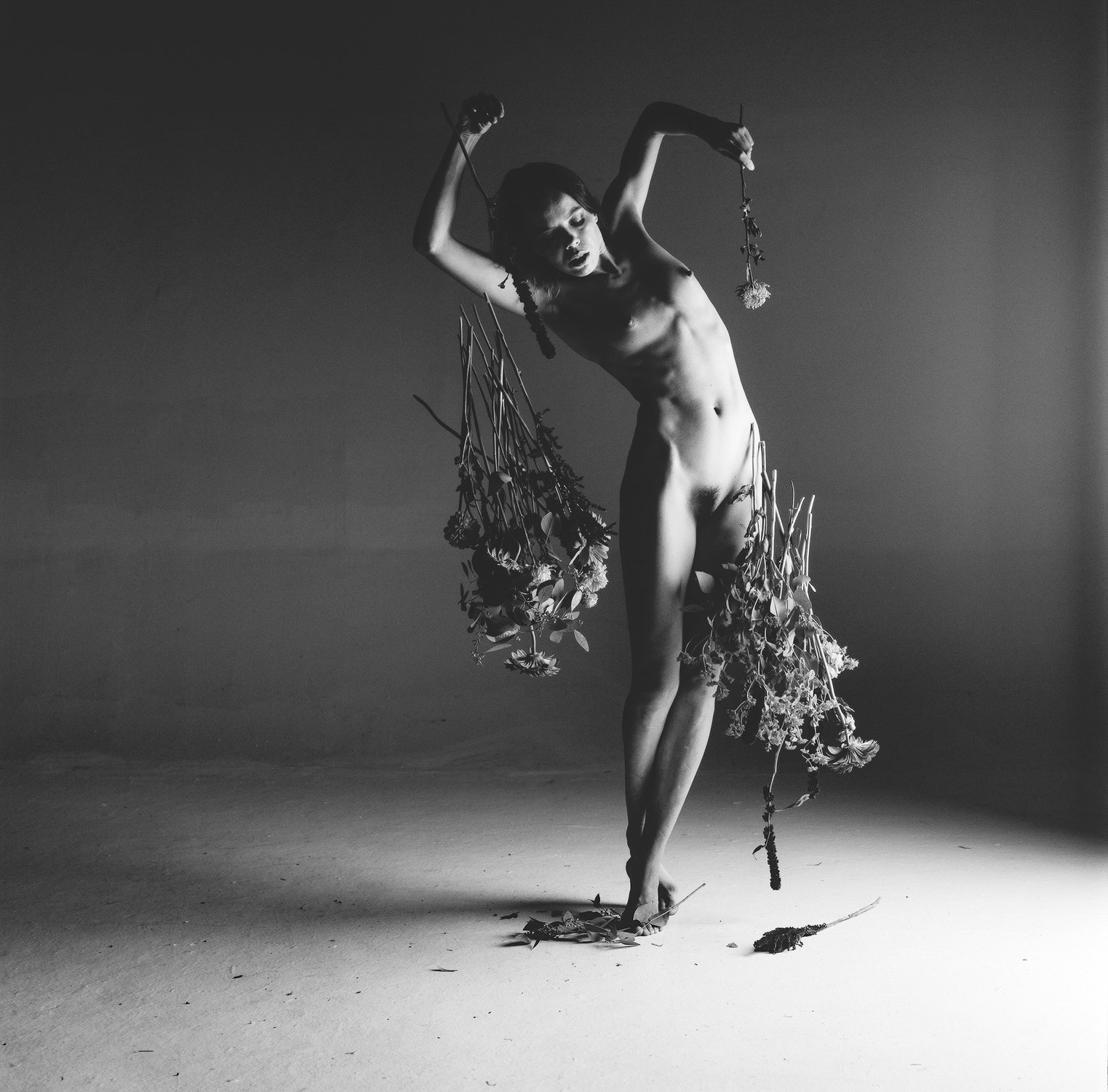 Black and white photo of a nude person holding flower bouquets, standing in a dramatic pose.