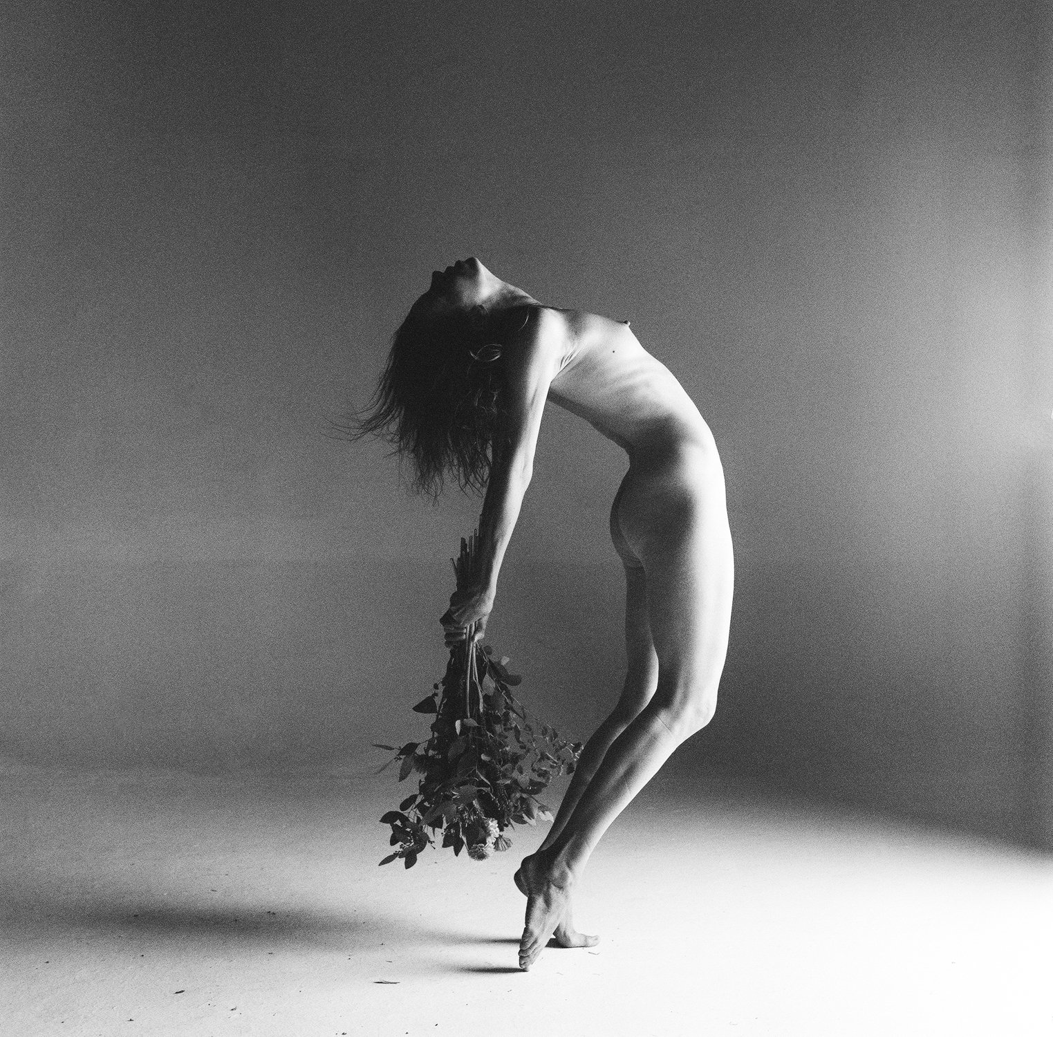 Black and white artistic photo of a nude person leaning back gracefully while holding a bouquet of flowers.