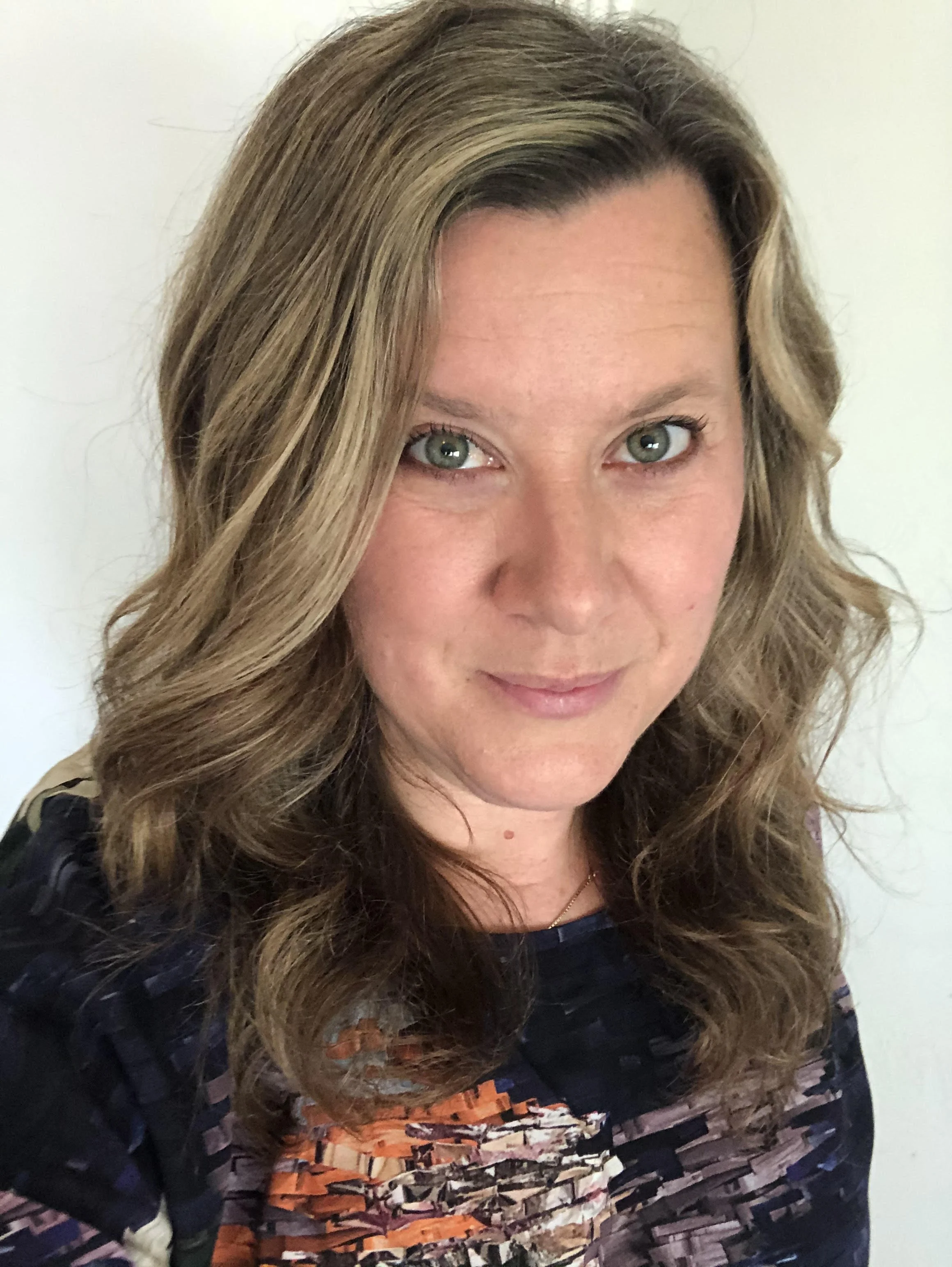 Close-up of a woman's face with wavy light brown hair with blonde highlights, blue eyes, and light makeup, wearing a patterned top with shades of orange, black, and purple, against a plain white background.