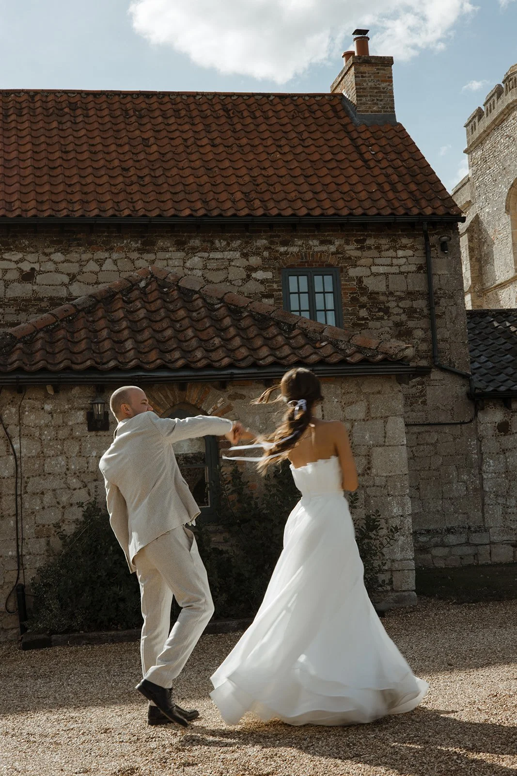A wedding couple, a man and a woman in a white dress, dancing outside a stone building with a red-tiled roof, in bright daylight.