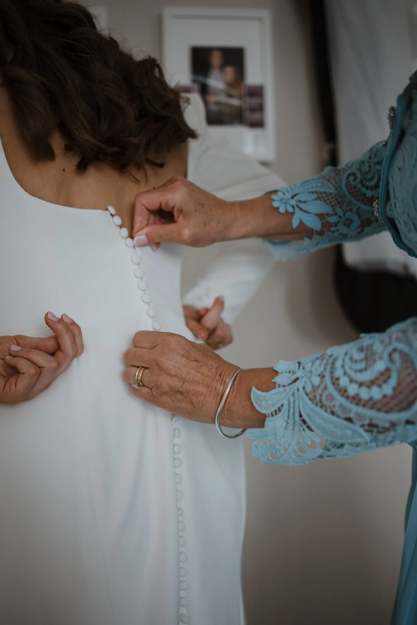 An elderly woman helps a bride button up the back of her white wedding dress.