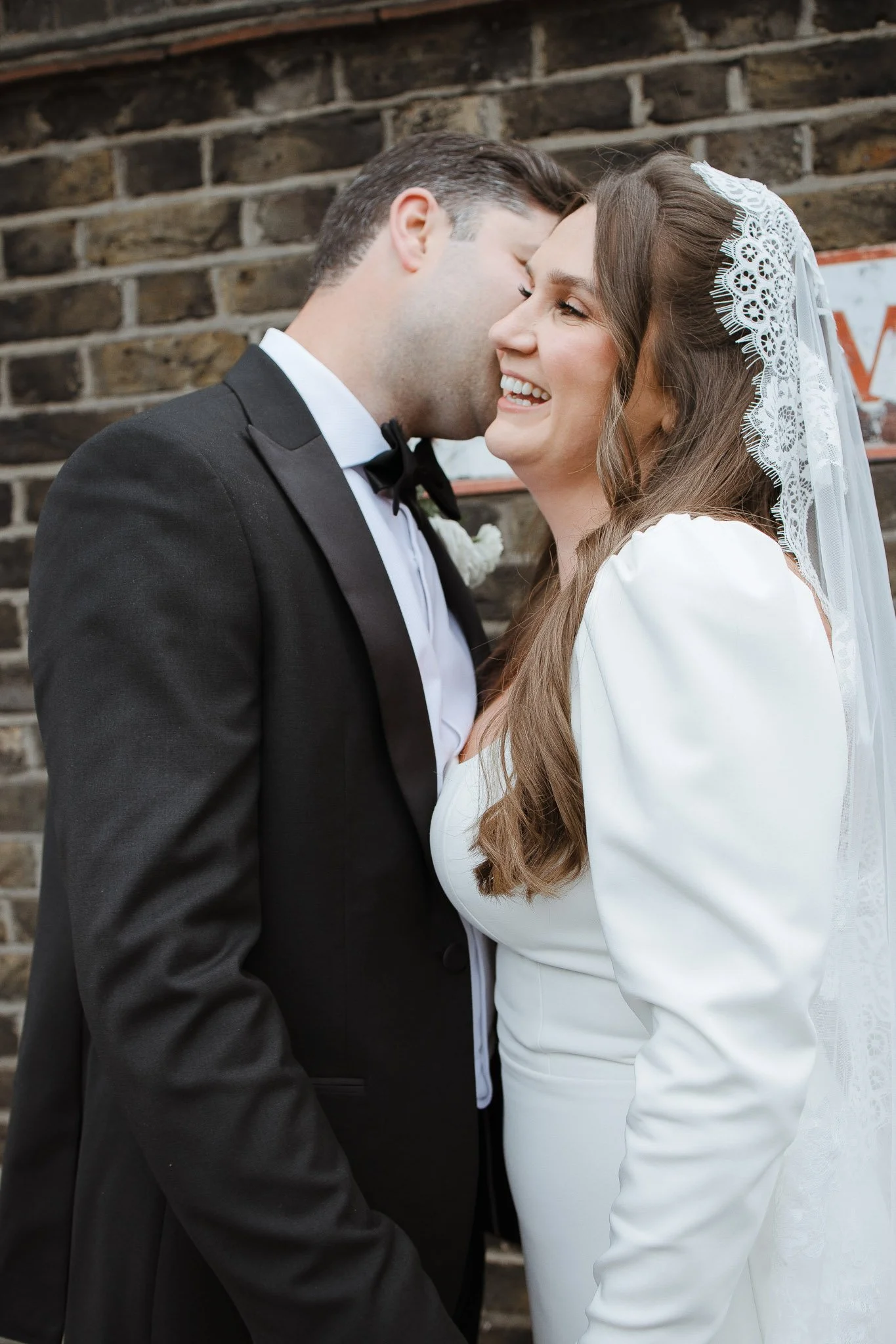 A bride and groom share a moment of laughter and affection, with the groom whispering into the bride's ear. The groom wears a black tuxedo and the bride wears a white wedding dress with lace trim and a veil. They are standing against a brick wall bac