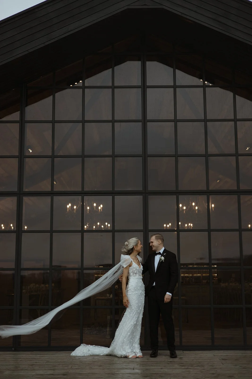 A bride and groom standing in front of a large glass window at sunset, gazing at each other. The bride is wearing a long white wedding dress with a veil, and the groom is dressed in a black tuxedo with a bow tie.