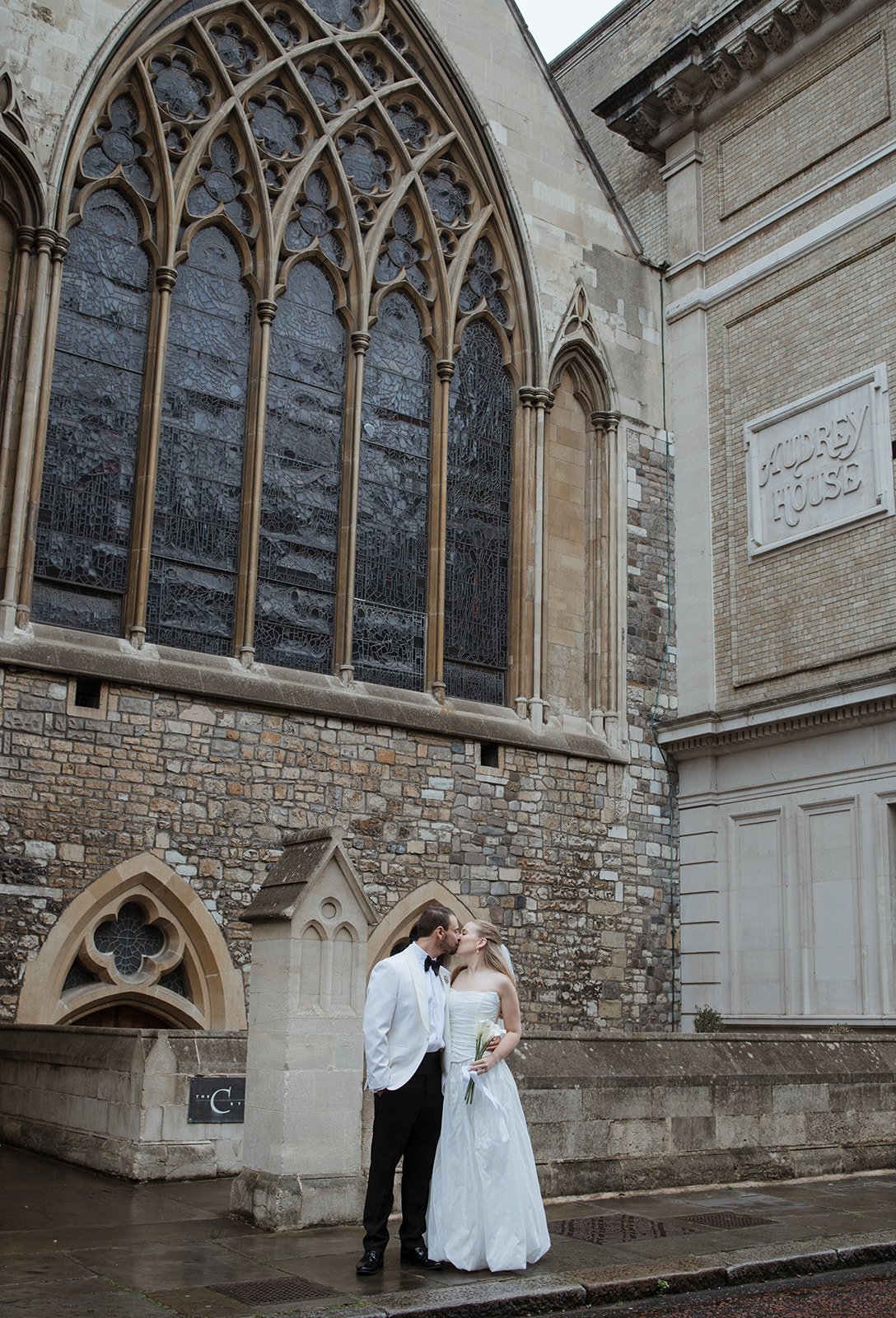 A bride and groom in wedding attire sharing a kiss outside a historic church with large stained glass windows.