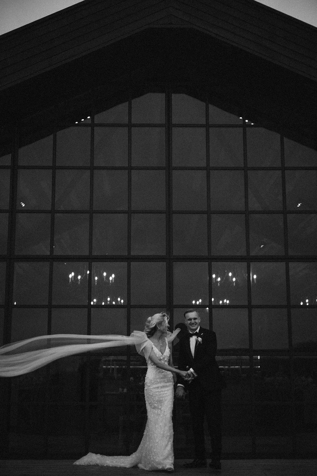 A black and white photo of a bride and groom holding hands in front of a large glass window with chandeliers reflected in it, inside a building with a peaked roof.