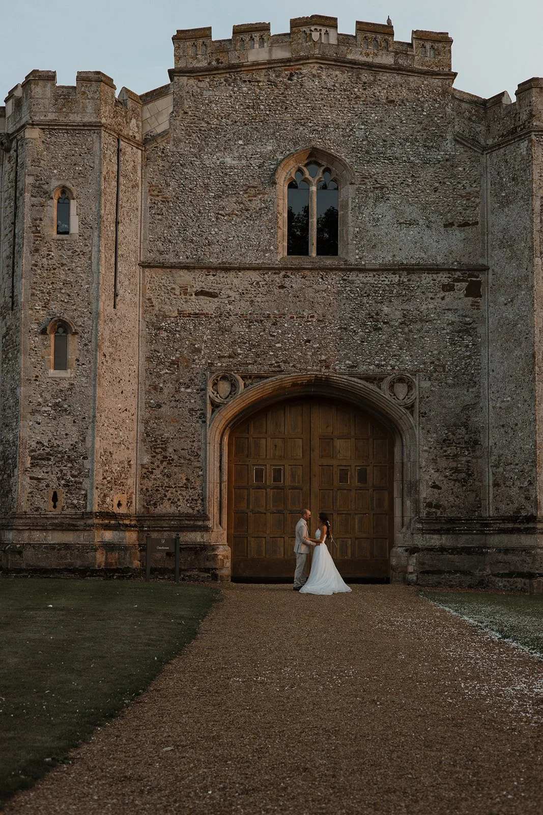 A bride and groom holding hands and facing each other in front of a large wooden door of an old stone castle or church at sunset.