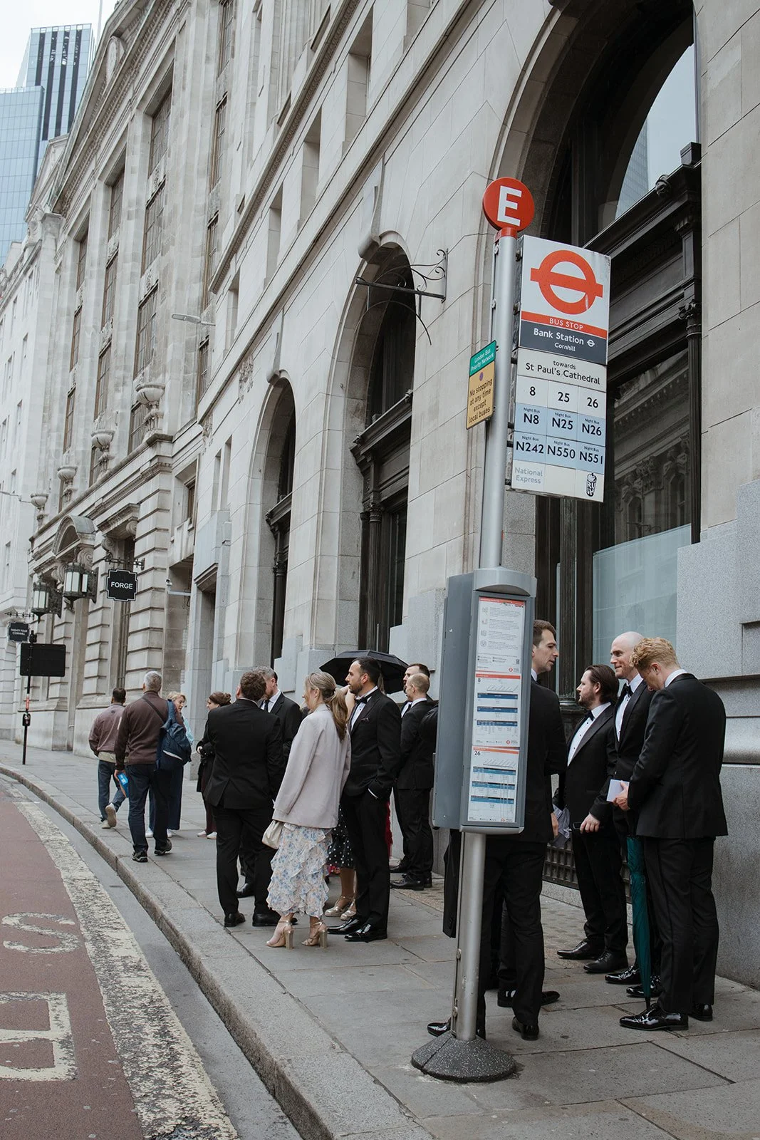 Group of people dressed in formal attire standing at a bus stop on a city street, with historic-looking buildings in the background.