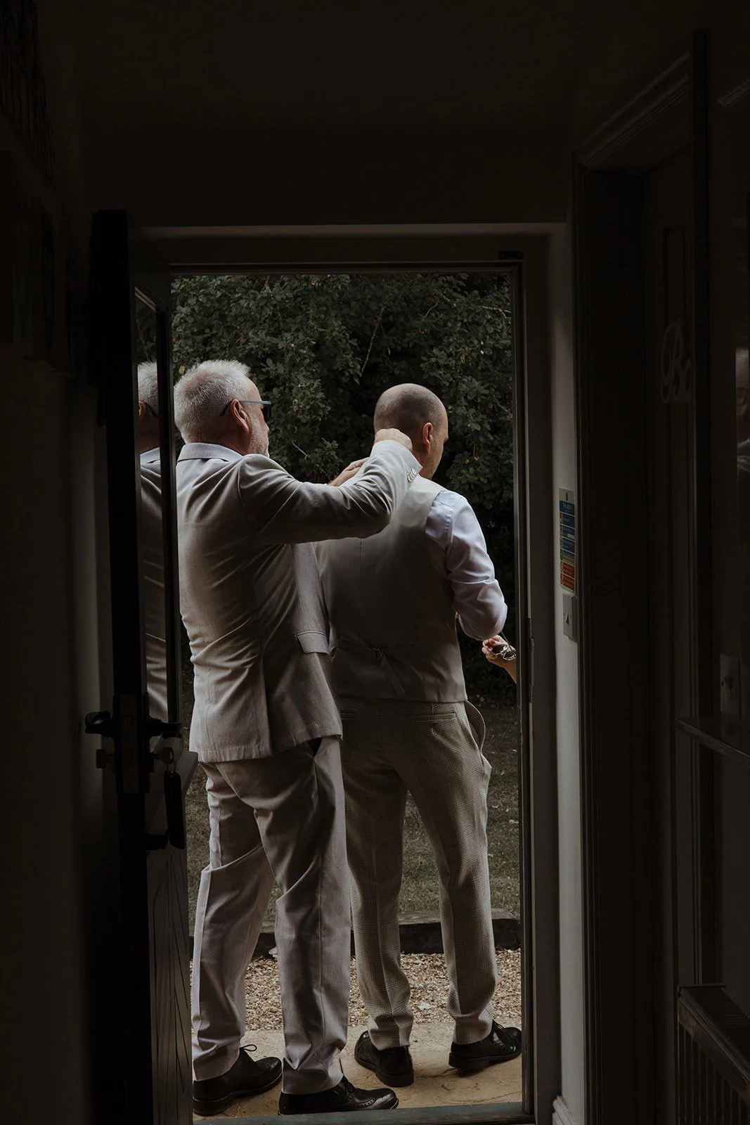 Two men in light-colored suits stand at the doorway, one helping the other with a haircut outside. The scene is viewed from inside a room with a dark interior, looking out onto greenery.
