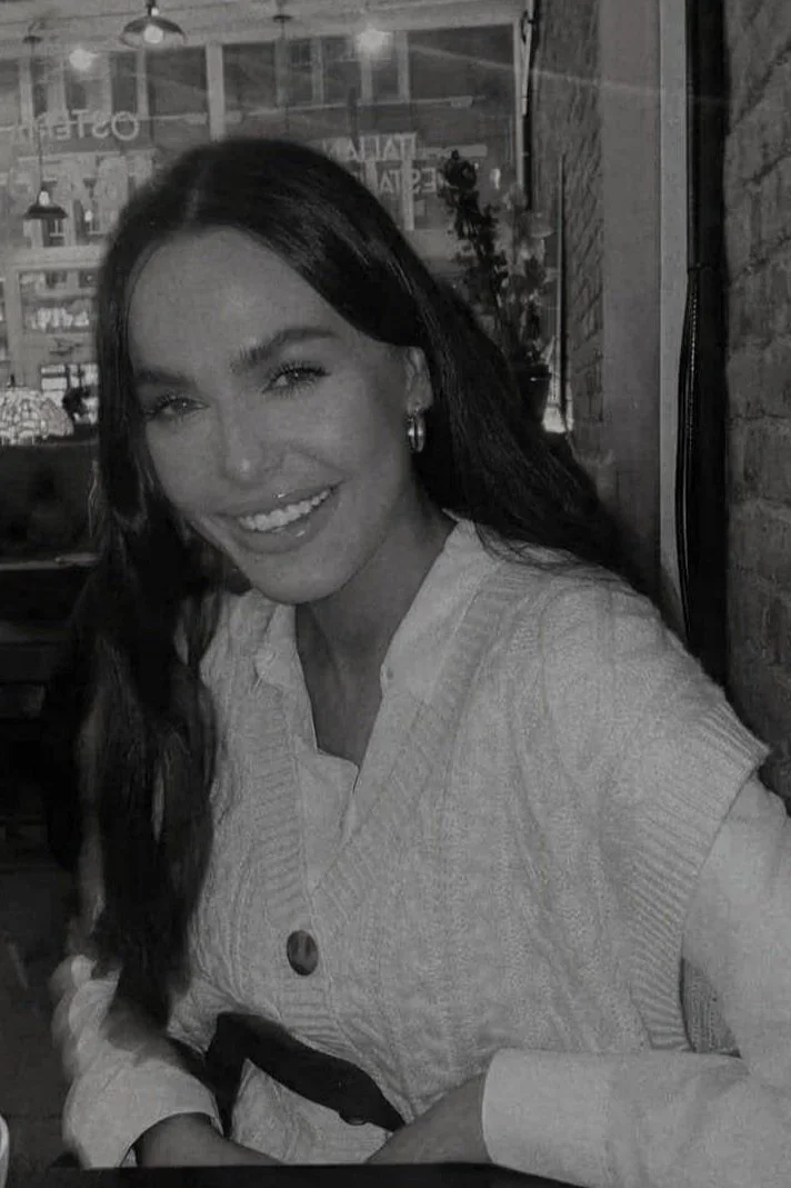 A smiling woman with long dark hair, wearing earrings and a light-colored sweater, sitting inside a restaurant or cafe with a window and brick wall in the background.