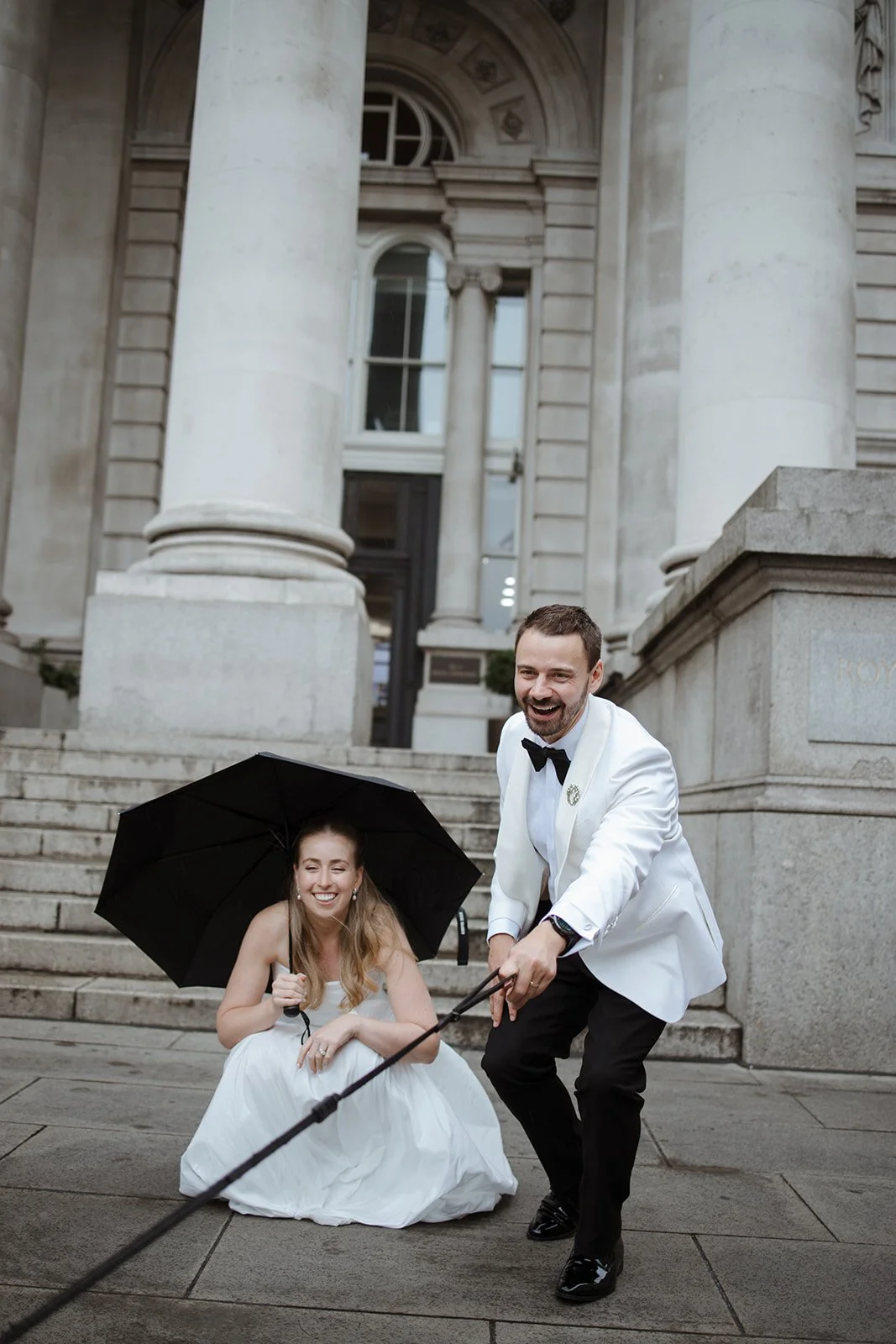 A man in a white tuxedo jacket and black bow tie pulls a woman in a white wedding dress on a leash while she sits on the steps holding an umbrella, both smiling and enjoying themselves outside a grand stone building with large columns.