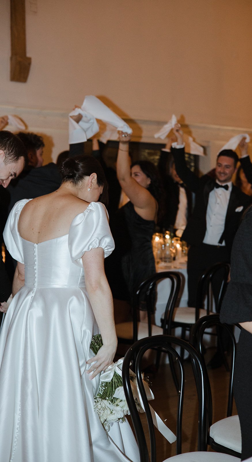 A bride in a white gown holding a bouquet with her head bowed, while guests in black attire raise napkins or cloths in celebration at a wedding reception.