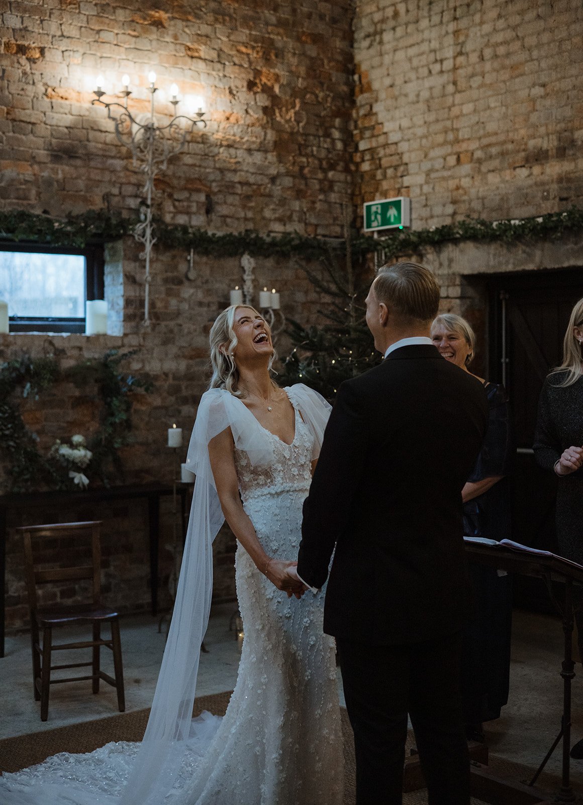 Bride and groom holding hands during wedding ceremony in rustic brick venue with candle decorations and a Christmas tree.