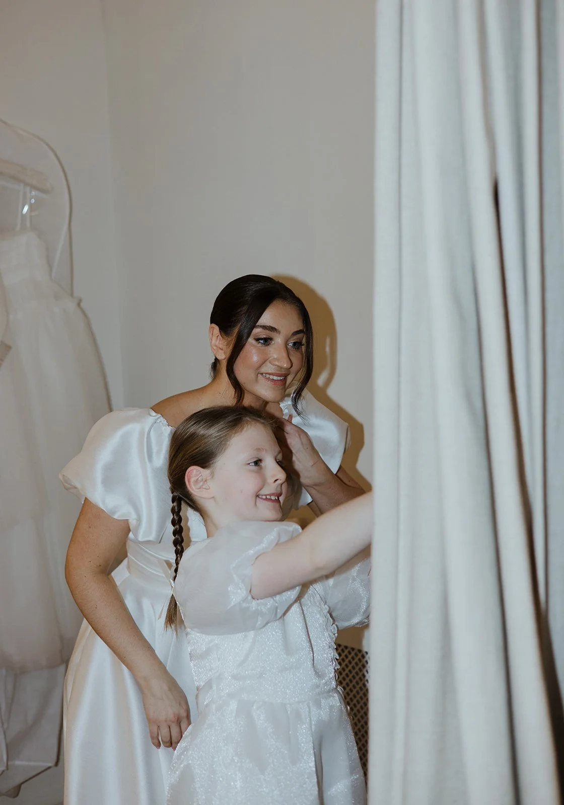 A woman and a young girl are trying on dresses in a fitting room, both wearing white dresses, with the woman helping the girl adjust her dress