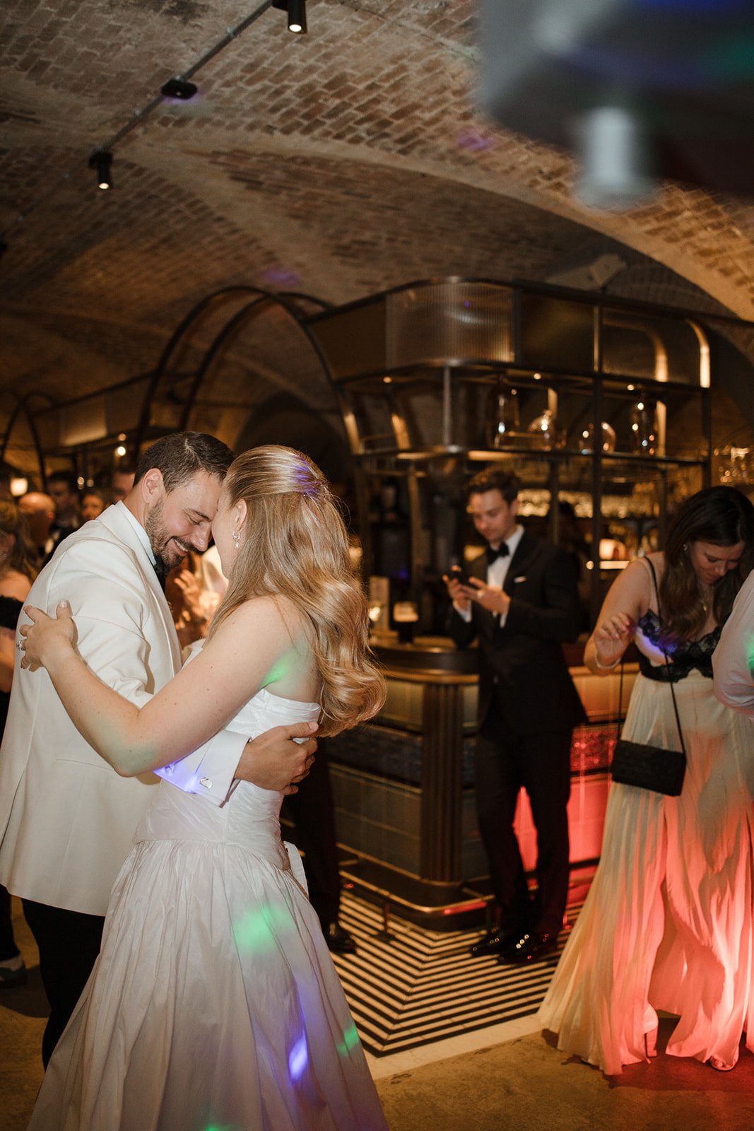Couple dancing at a wedding reception in a stylish venue with arched brick ceilings, surrounded by other guests, some dancing and others socializing.