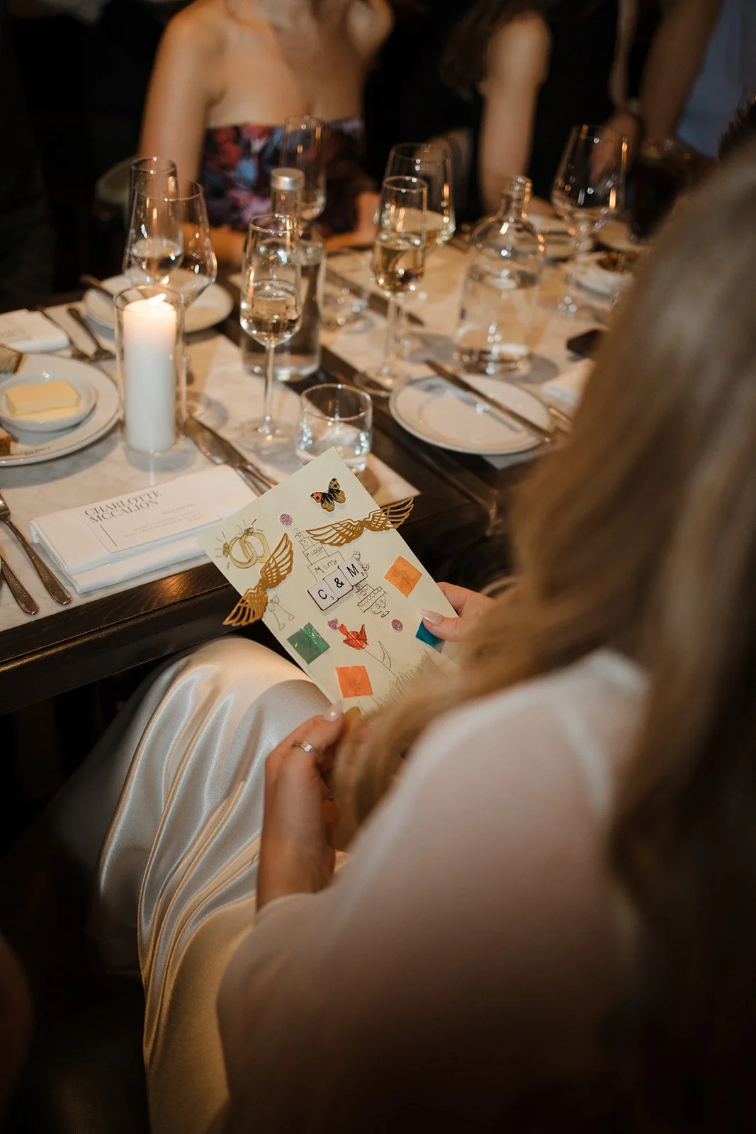 Person holding a colorful card with butterfly and wing designs at a formal dinner table set with multiple wine glasses, plates, and candles.